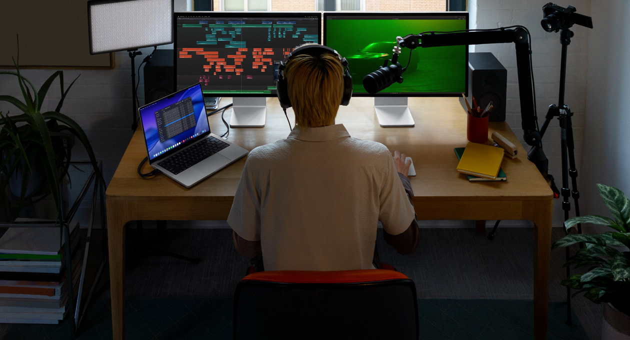 Persona sentada en un escritorio con un micrófono y audífonos frente a tres dispositivos: un monitor Studio Display para la Mac, herramientas de edición de audio, clips de sonido en diferentes bloques de color, un segundo monitor Studio Display para la Mac, foto de un auto en la niebla con iluminación ambiental verde, y una MacBook Pro de 16 pulgadas con la app Finder abierta, Archivos para Final Cut Pro, el Dock y un fondo de escritorio azul estilizado