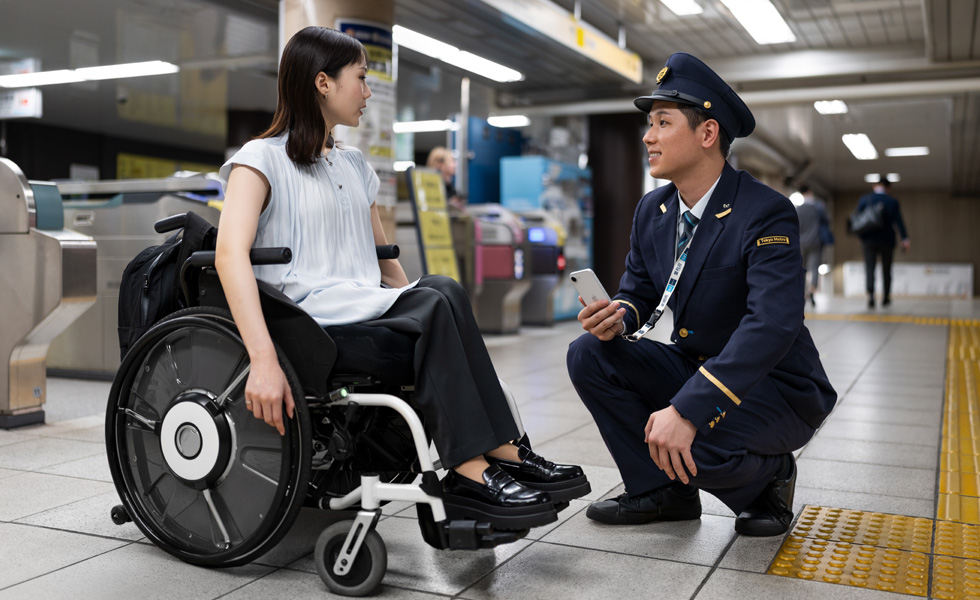 Một phụ nữ ăn mặc lịch sự ngồi trên xe lăn đang trao đổi với một nhân viên ga tàu ở sân ga.